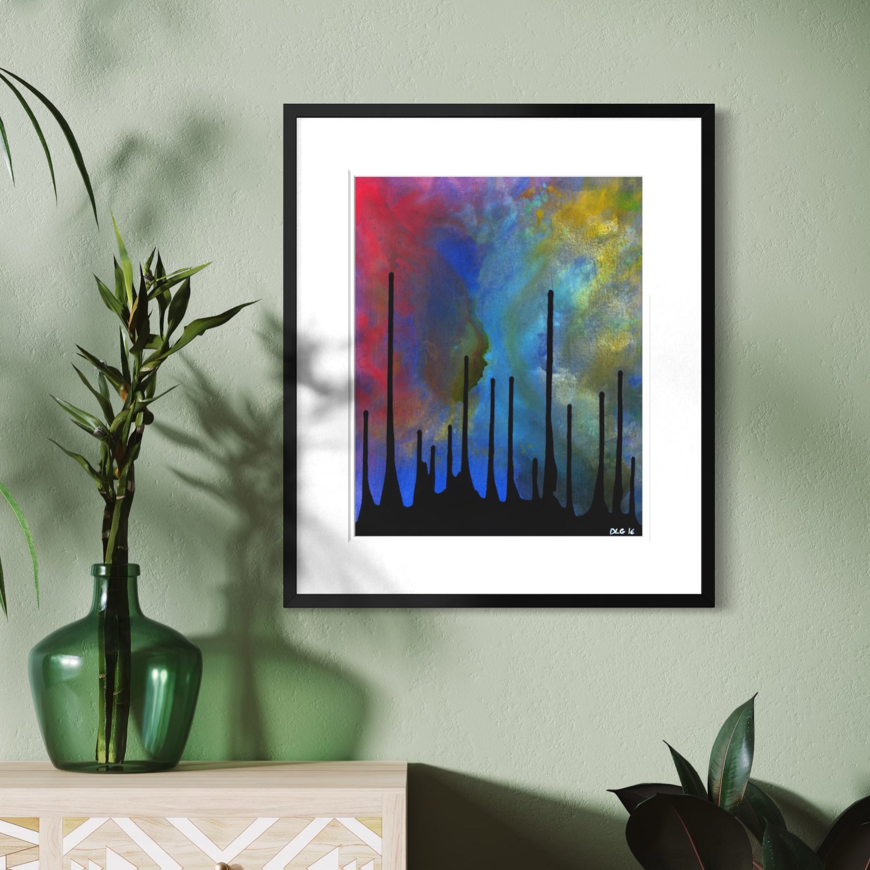 A framed abstract art print featuring vibrant blue, red, yellow, and green brushstrokes with black vertical silhouettes, displayed on a sage green wall above a light wood console table with a green glass vase.